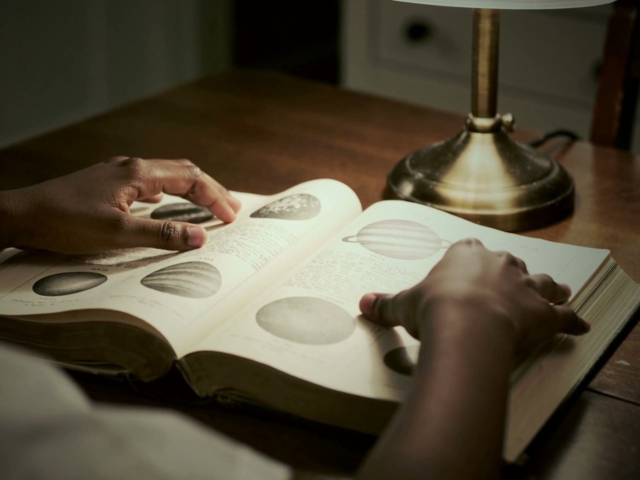 Hands turning the pages of an old astronomy book under a desk lamp, a scene from the film